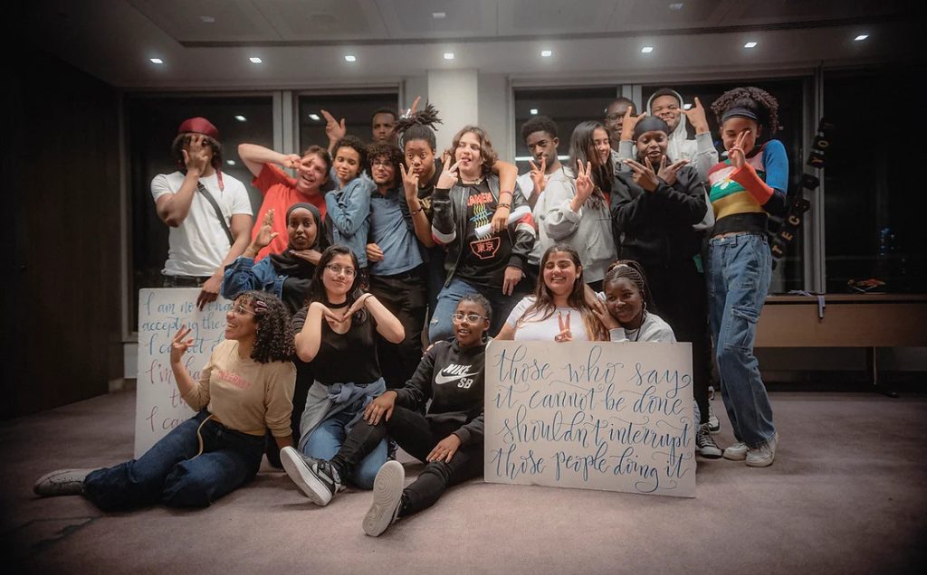 Group of young people, who are part of the Advocacy Academy, smiling and posing for the photo. They have a sign which says 'those who say it cannot be done shouldn't interrupt those people doing it'