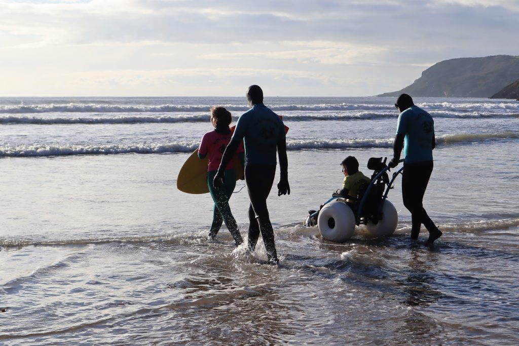 A child in a wheelchair going into the sea with three adult surfers in a project run by Surfability
