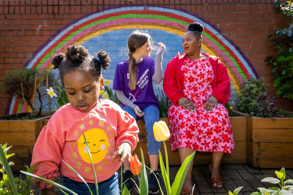 A child playing in a garden. In the background, a member of the Home Start team is talking to the child's carer.