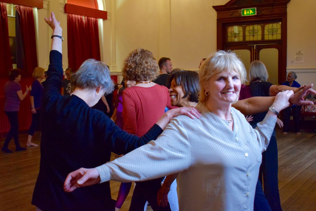 Older people dancing in rehearsal for a production. They are joining arms and smiling.